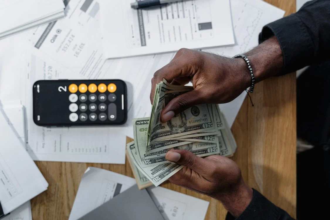 An image of a person counting currency bills with documents and a calculator lying around as they wonder what investments they should make.