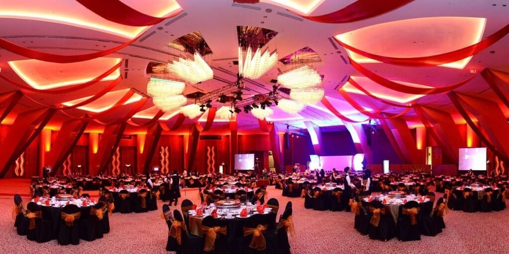 alt= A large, elegantly decorated ballroom features round tables with black and gold table settings, under a ceiling with ornate chandeliers and red, draped accents.