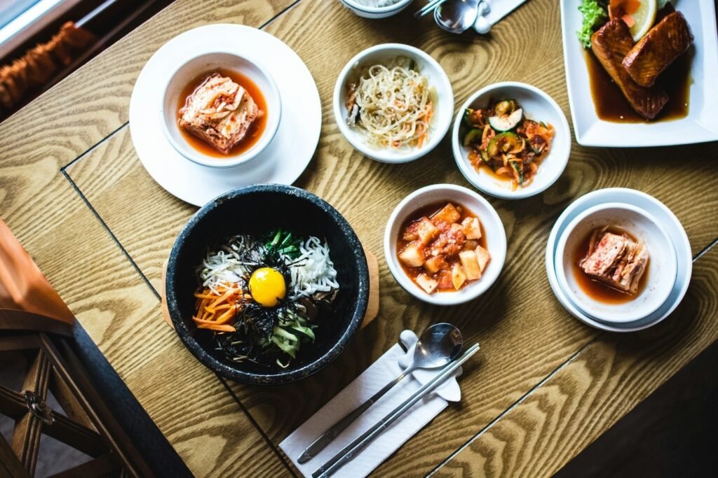 A traditional Korean meal featuring bibimbap in a stone bowl with a raw egg yolk on top, surrounded by various banchan (side dishes) including kimchi, pickled vegetables, japchae (stir-fried glass noodles), and grilled fish, served on a wooden table.