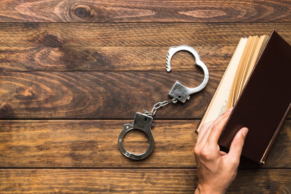 Handcuffs lie open on a wooden surface next to a hand holding an open book.