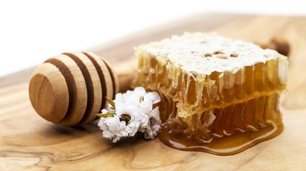 lose-up of fresh manuka honeycomb oozing golden honey on a wooden board, with a wooden honey dipper and a white leptospermum flower beside it.